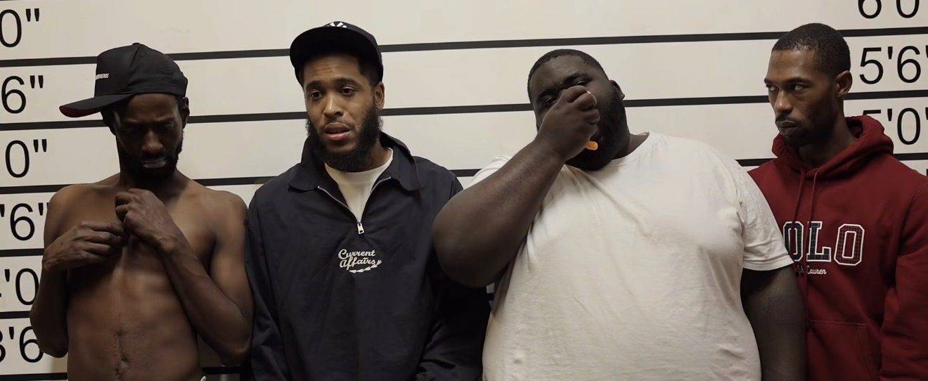 Still from a comedy sketch: four characters lined up against a police-lineup height chart, each reacting differently to the camera.