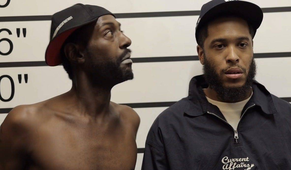 Still from a comedy sketch: two characters standing against a police-lineup height chart, one looking sideways at the other.