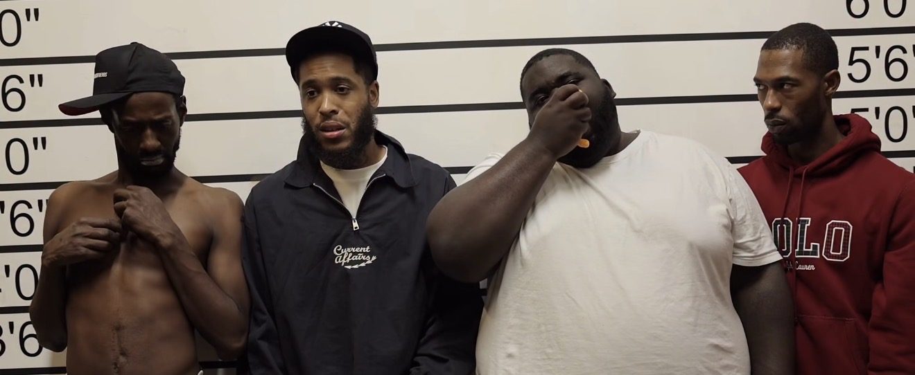 Still from a comedy sketch: four characters lined up against a police-lineup height chart, each reacting differently to the camera.