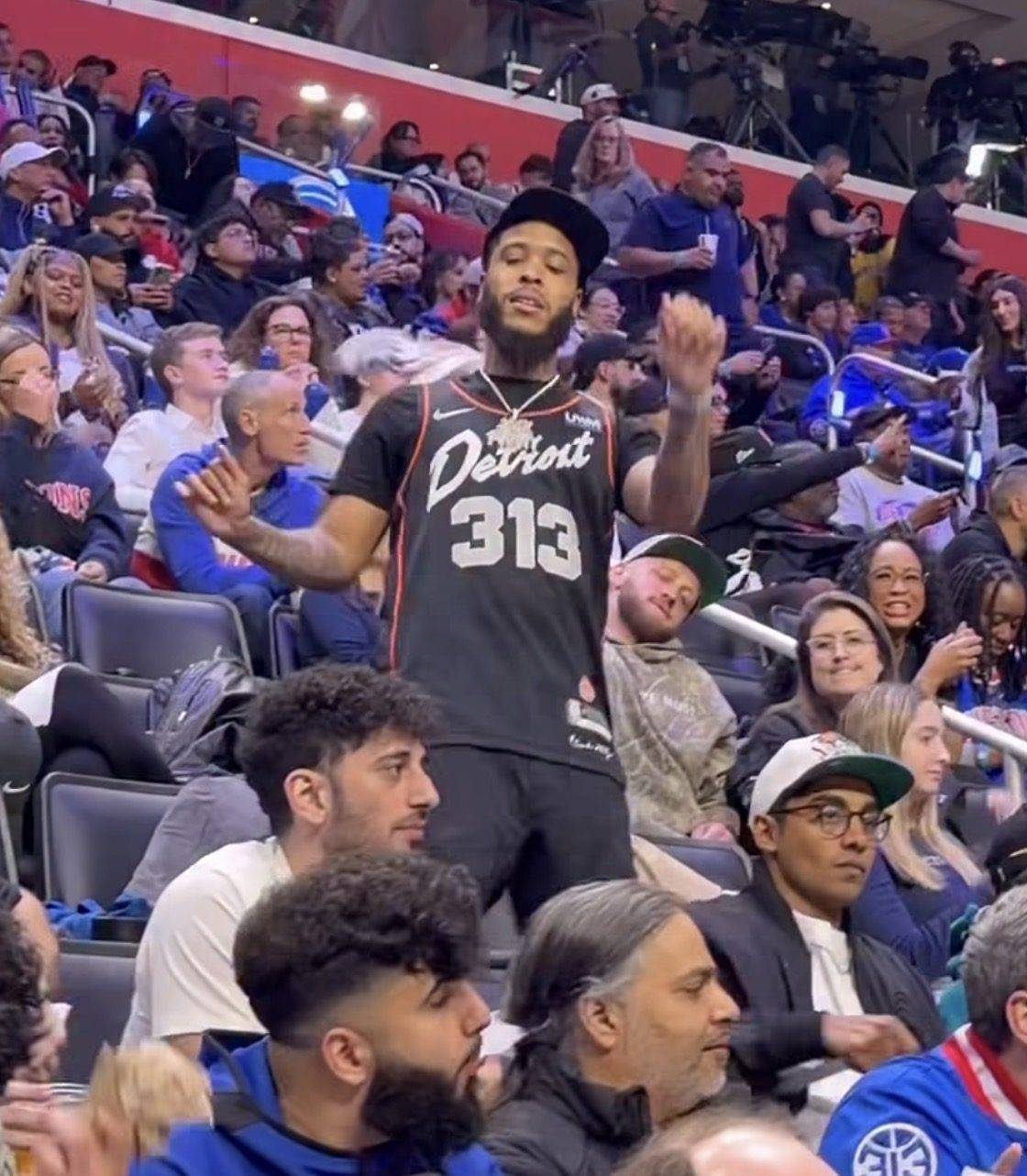 Jack Funny standing in the stands at a Detroit basketball game, wearing a black Detroit 313 jersey and a diamond chain.